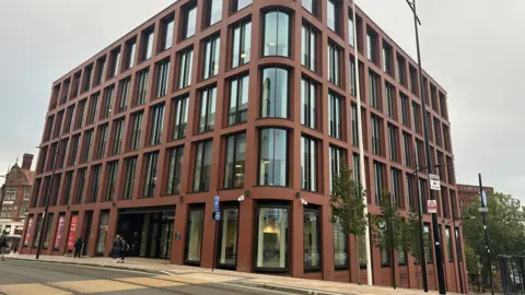 A modern five-story brick building with large windows, including curved windows on the corner facing the camera. 