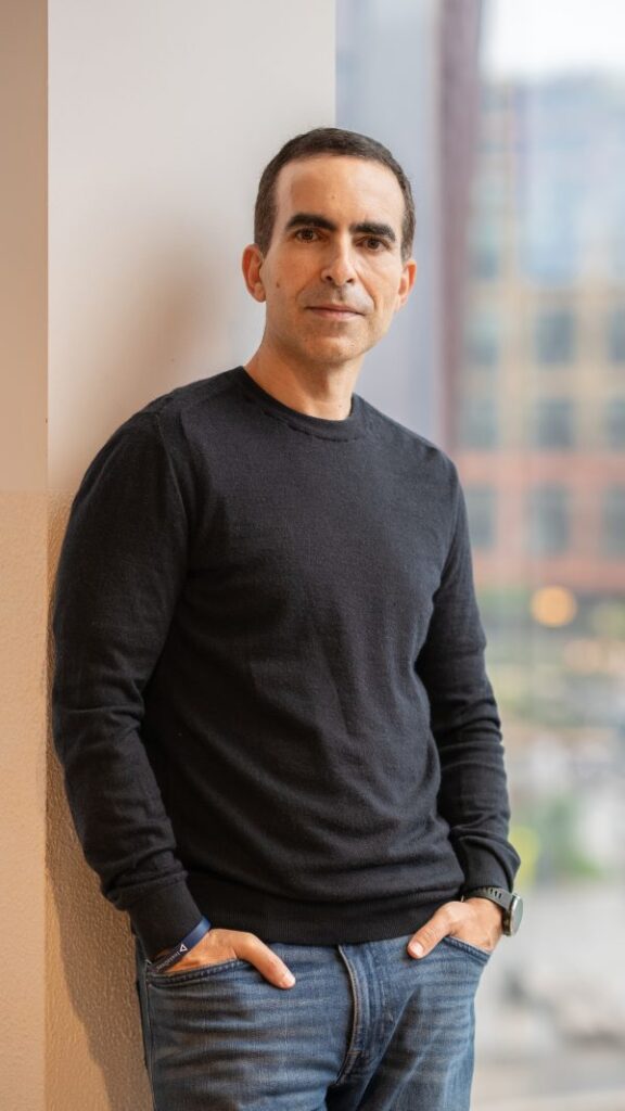 Portrait of Karim Beguir, wearing a black long-sleeved T-shirt and jeans, with his hands in his pockets, leaning against a wall, next to a large window.