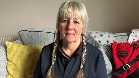 BBC Susan O Sullivan, with blonde braids in her hair and wearing a black top, sits on a grey sofa with a yellow cushion.